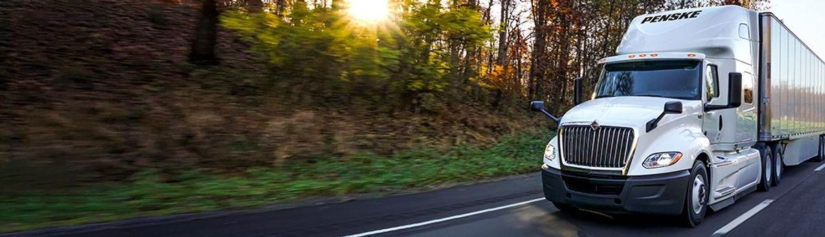 A white Penske semi-truck drives on a road at sunrise.