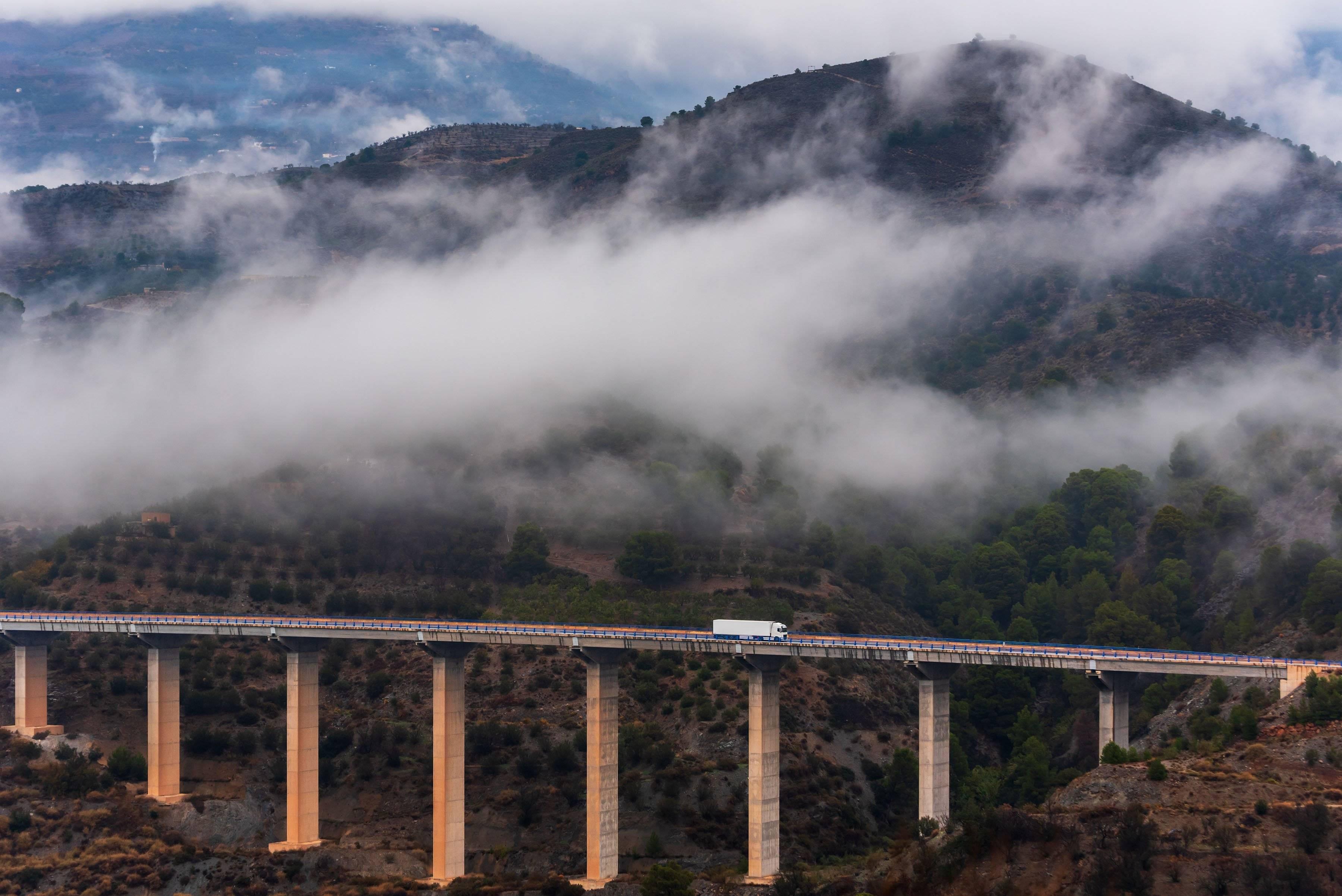 A white semi-truck drives across a bridge through foggy mountains.