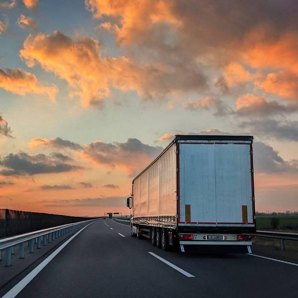 A white semi-truck drives away from the camera under an orange sunset sky.