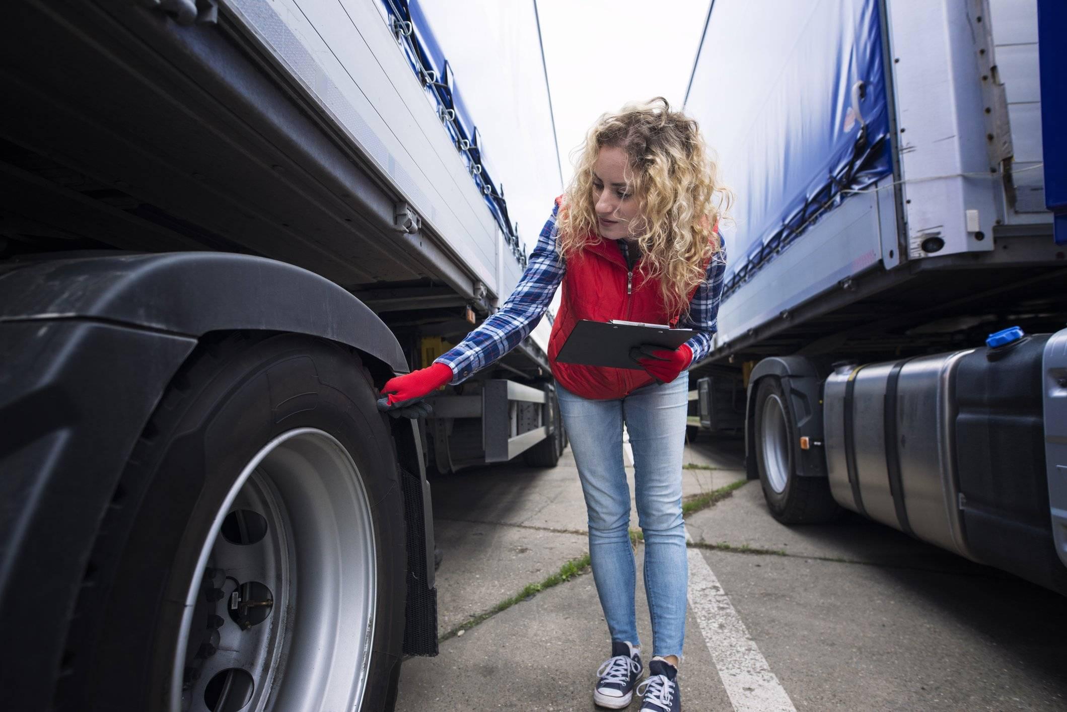 A woman holds a clipboard and checks the tire of a semi-truck.
