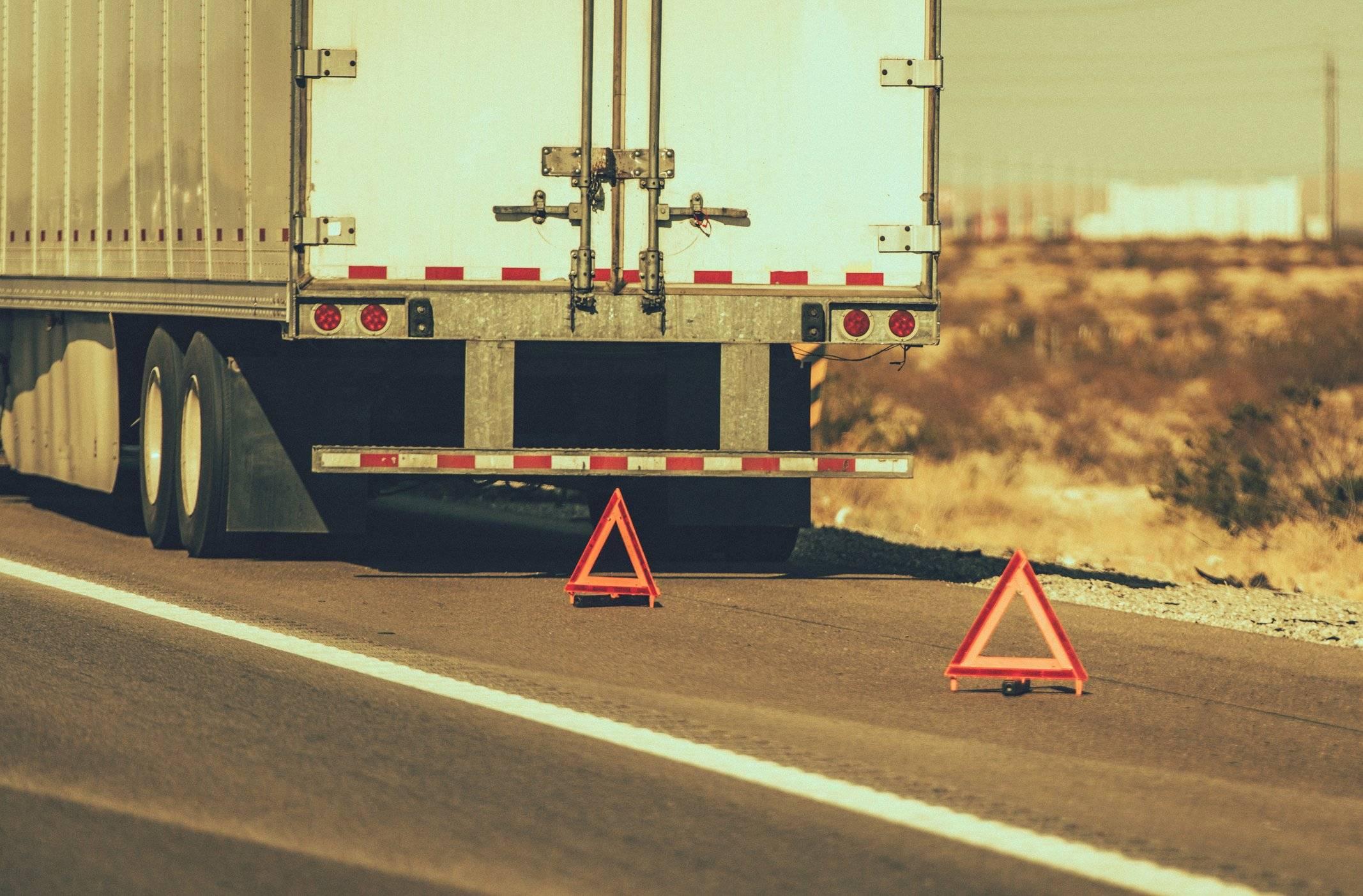 The back of a white semi-truck parked on the side of a road with two caution triangles set up behind it.